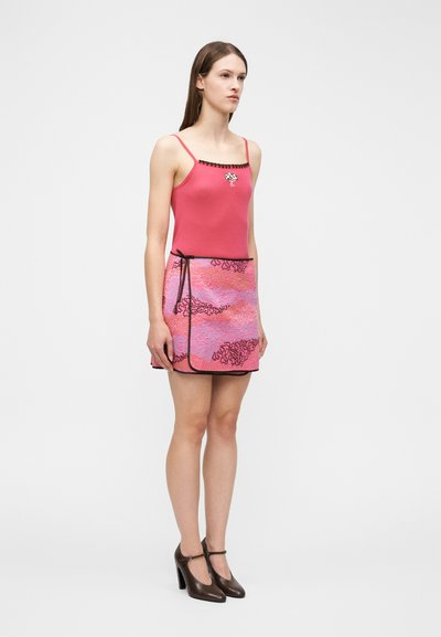 Γυναίκα που στέκεται, φορώντας ροζ τοπ με λεπτές τιράντες και μαύρη μπορντούρα, ροζ φούστα τύλιγμα με μοτίβα σε μωβ, και μαύρα παπούτσια με ψηλό τακούνι.