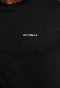 Černé bavlněné tričko s kulatým výstřihem a malým bílým logem "ARMANI EXCHANGE" na levé části hrudi. Hladká textura, přiléhavý design.