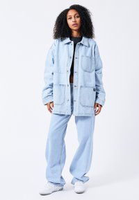 Light blue denim jacket with pockets, button closure, and loose fit, paired with matching wide-leg pants and white sneakers.