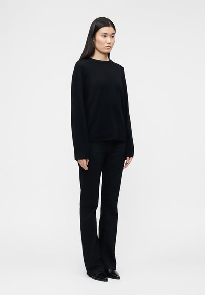 Γυναίκα όρθια, φοράει μαύρο μακρυμάνικο τοπ, μαύρο παντελόνι και μαύρα μυτερά παπούτσια, μπροστά σε απλό ανοιχτό γκρι φόντο.