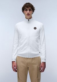 Vit zip-up jacka med hög krage, strukturerad stickad design och en cirkulär logotyp patch. I kombination med ljusbeige cargobyxor med fickor.
