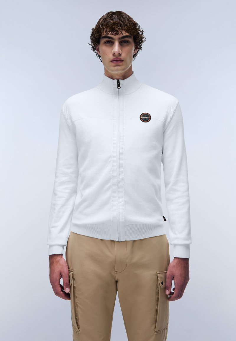 Vit zip-up jacka med hög krage, strukturerad stickad design och en cirkulär logotyp patch. I kombination med ljusbeige cargobyxor med fickor.