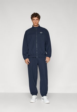 Navy tracksuit made of lightweight fabric, featuring a zip-up jacket with elastic cuffs and matching pants with elastic hems. White sneakers.
