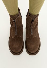 Stivaletti in suede marrone con punta arrotondata, zip laterali e una finitura testurizzata, abbinati a calzettoni a coste di un verde chiaro.