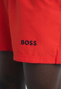 Raudoni šortai, pagaminti iš lygios medžiagos, pasižymintys tamsiai mėlynu "BOSS" logotipu apatinėje dalyje ir standartine apdaila. Atrodo, kad yra laisvi.