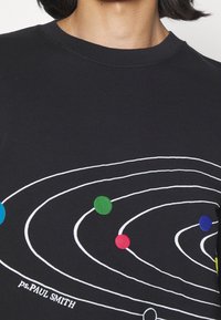 T-shirt noir à col montant avec un design coloré du système solaire, comprenant des lignes d'orbite et des points représentant les planètes, accompagné du texte "ps.PAUL SMITH" près de l'ourlet.