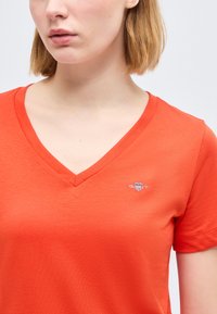 Νεαρή γυναίκα που φοράει πορτοκαλί απλό T-shirt με V λαιμόκοψη και μικρό λευκό κεντητό λογότυπο στο αριστερό στήθος.