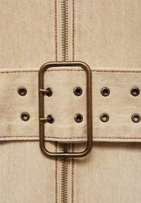 Tissu en denim beige avec une fermeture éclair en métal et une boucle rectangulaire en bronze. Comprend des œillets décoratifs le long de la ceinture et des coutures contrastantes.