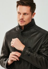 Zwarte leren jas met een hoge kraag, voorzien van grote knopen en een gestructureerde afwerking. De mouwen hebben knoopaccenten. Close-up weergave.