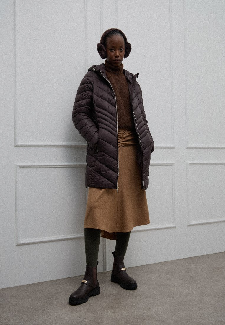 Brązowa kurtka puchowa, długie rękawy, zamek błyskawiczny. Brązowy sweter, beżowa spódnica, zielone rajstopy. Czarne botki za kostkę z złotymi akcentami. Neutralne tło.