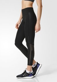 Nainen, jolla on yllään mustat korkeavyötäröiset legginsit, joissa on verkkopaneelit sääriin, ja mustat Adidasin juoksukengät, joissa on valkoiset pohjat.