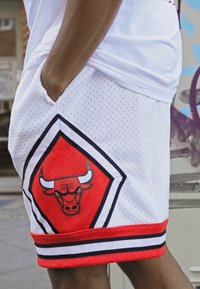 Pantalones cortos de baloncesto de malla blanca con acentos rojos, que presentan un gran logotipo de los Bulls en rojo y negro en el lado izquierdo y un dobladillo a rayas en negro y rojo.