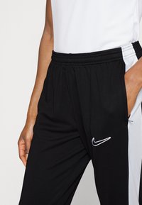 Zwarte sportbroek met witte zijkanten, elastische tailleband en schuine zakken. Heeft een wit Nike-logo op de linker bovenbeen.