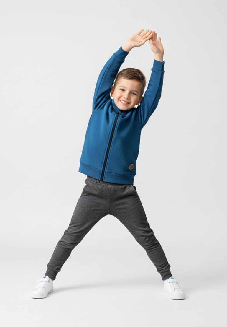 Niebieska kurtka na zamek z wysokim kołnierzem, szare spodnie dresowe i białe trampki. Strój ma małe logo na kurtce.