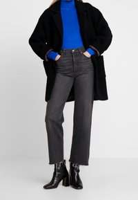 Schwarzer strukturierter Mantel über einem blauen Rollkragenpullover, kombiniert mit hoch taillierten grauen Jeans und schwarzen Ankle Boots. Auffälliger Fransenabschluss an den Jeans.