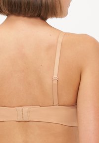 Nude bra featuring smooth fabric, adjustable straps, and a hook-and-eye clasp at the back. Minimalist design with no visible embellishments.