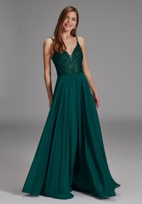 Jeune femme portant une robe sans manches vert foncé avec un corsage à sequins et une jupe fluide, debout et souriante devant un fond gris.