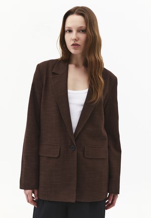 Jonge vrouw met lang bruin haar draagt een oversized donkerbruine blazer over een witte top, staand tegen een effen witte achtergrond.