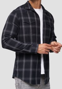 Czarna i szara koszula flanelowa w kratę z długimi rękawami, zapinana na guziki z dwoma kieszeniami na piersi, noszona na białym t-shircie.