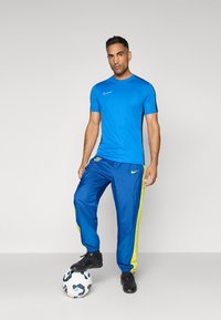 Niebieska koszulka sportowa z krótkim rękawem i czarnymi akcentami, połączona z niebieskimi spodniami dresowymi z żółtymi paskami, czarnym obuwiem i piłką nożną.