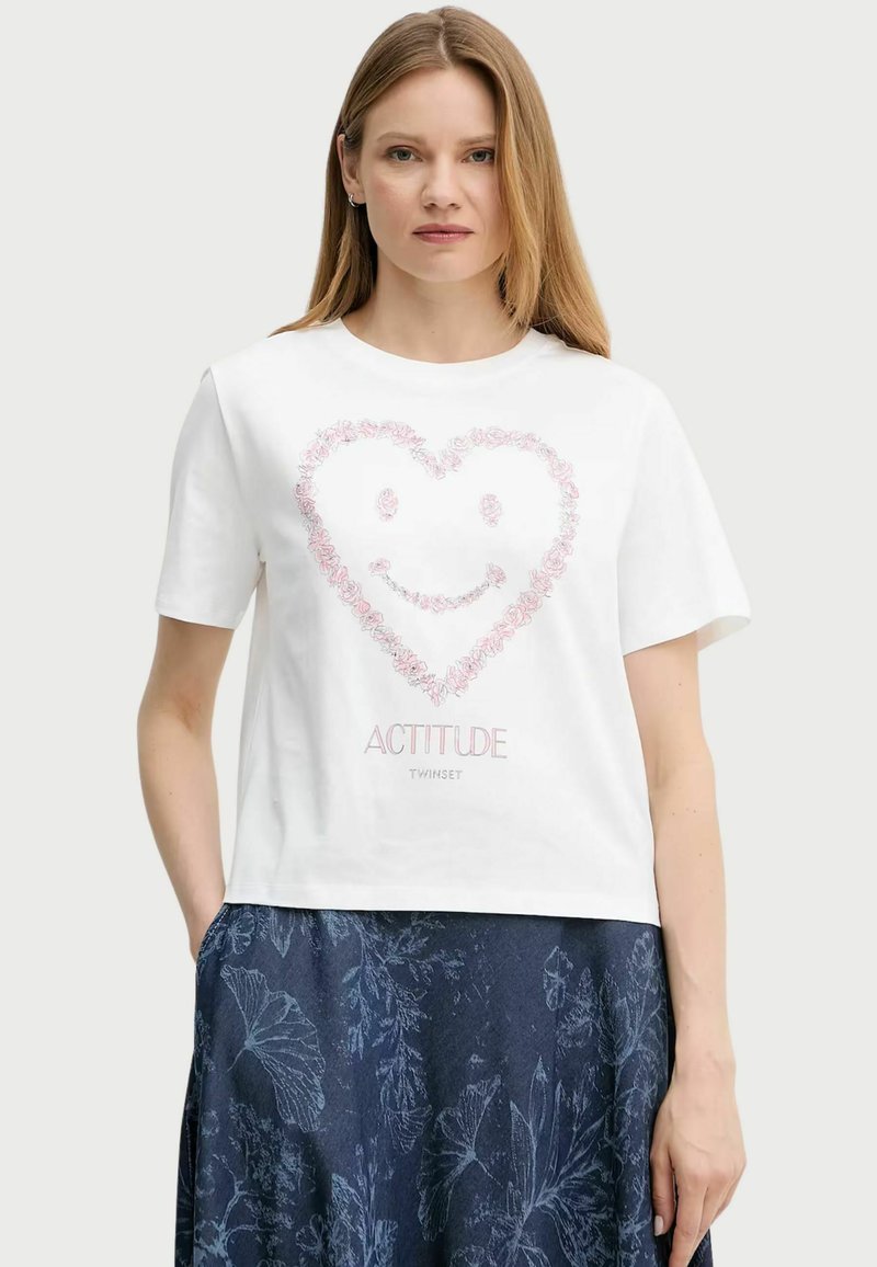 T-shirt bianca in cotone con maniche corte, caratterizzata da un design a forma di cuore floreale rosa e una faccina sorridente, abbinata a una gonna blu a fantasia.