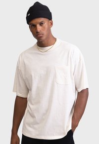 Relaxed-fit, light beige t-shirt with short sleeves, featuring a chest pocket. Smooth texture, cotton material, and a crew neckline.
