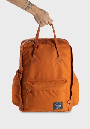 Mochila de tela naranja con un bolsillo frontal y bolsillos laterales de malla. Cuenta con asas resistentes y un parche con el logotipo. Material liso y con textura.