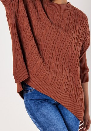Personne portant un pull en maille torsadée couleur rouille avec un ourlet asymétrique et un jean bleu, posant avec un bras levé.