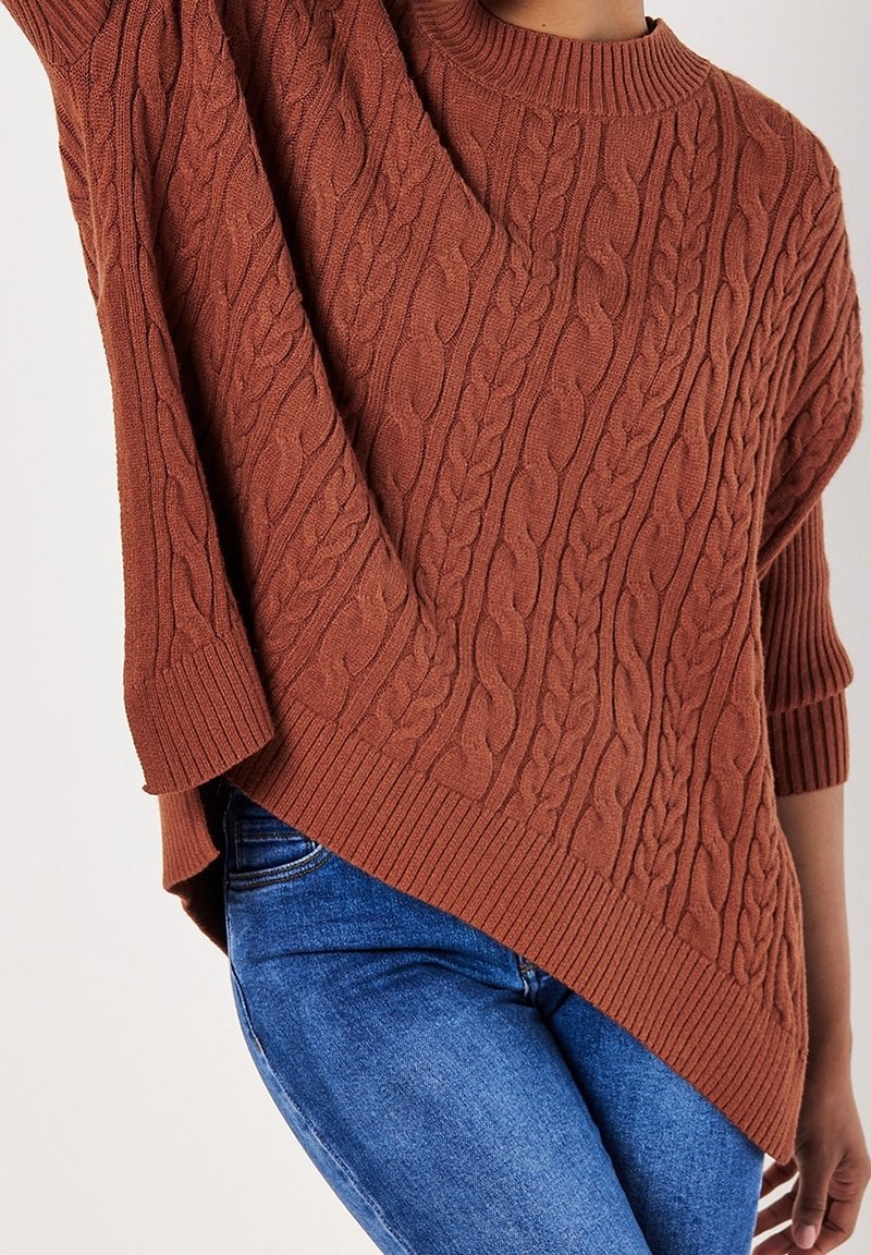 Personne portant un pull en maille torsadée couleur rouille avec un ourlet asymétrique et un jean bleu, posant avec un bras levé.