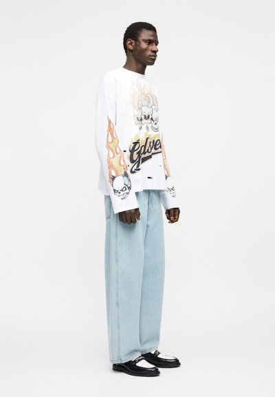 Νεαρός άνδρας στέκεται, φοράει άσπρο μπλουζάκι μακρυμάνικο με γραφικά καυτών κρανίων και φαρδιά ανοιχτό μπλε τζιν με μαύρα και λευκά παπούτσια.