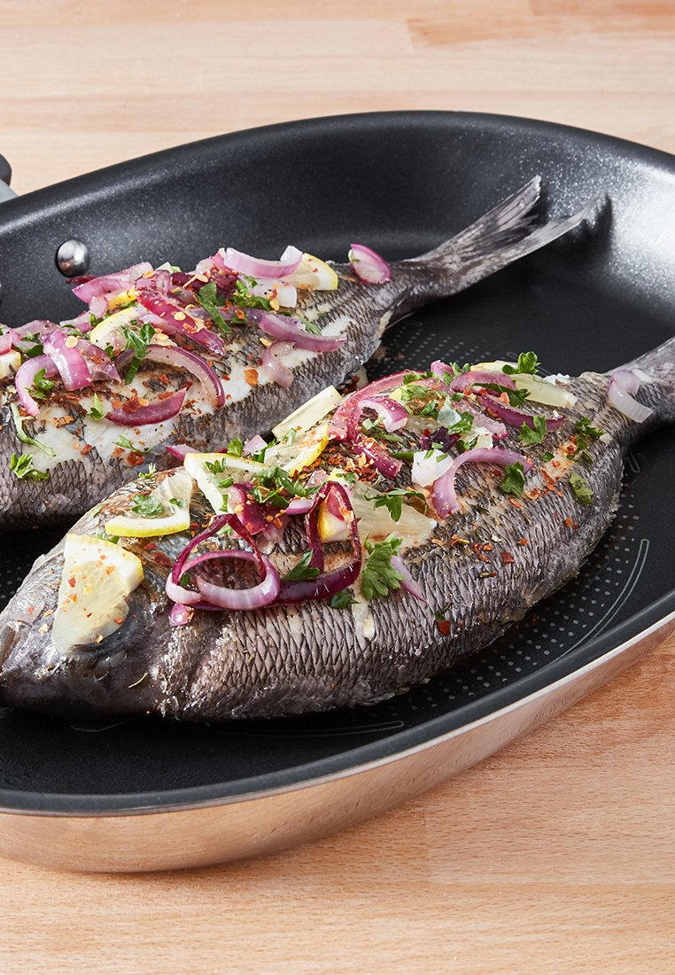 Cała ryba na czarnej patelni, ozdobiona plasterkami czerwonej cebuli, zielonymi ziołami, cytryną i przyprawami; skóra jest ciemnoszara z teksturą łusek.