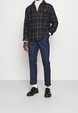 Mies, joka pukeutunut laivastonsiniseen ruudulliseen takkiin valkoisen pooloneuleen päällä, laivastonsinisiin räätälöityihin housuihin, valkoisiin sukkiin ja mustiin nahkakenkiin, seisomassa.