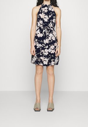 Femme portant une robe sans manches bleu marine à imprimé floral rose, avec une taille nouée, et des sandales à talons blocs ouvertes beige, debout devant un fond uni.