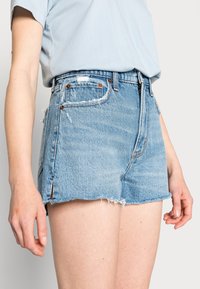 Persona vistiendo pantalones cortos de mezclilla de alta cintura en azul claro con detalles desgastados y una camiseta metida de color azul pálido, de pie contra un fondo liso.