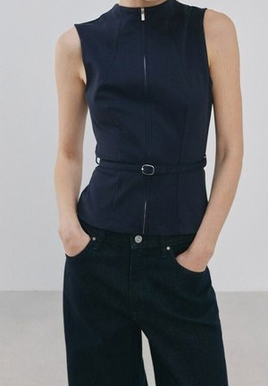 Personne portant un top noir sans manches avec fermeture éclair à l'avant et ceinture fine, associé à un pantalon noir taille haute, mains dans les poches.
