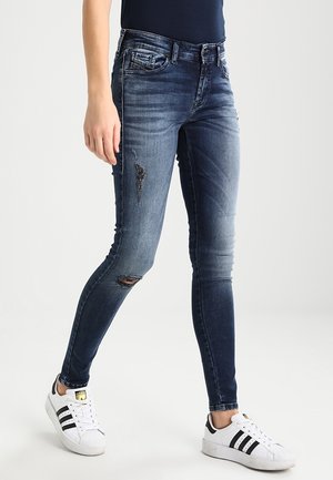 Jeans skinny en denim bleu avec des détails délavés et des déchirures, accompagnés de baskets blanches. Les jeans présentent un design classique à cinq poches.