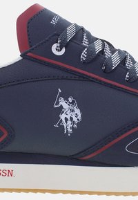 Tengerészkék sneaker sima szintetikus felsőrésszel, piros kiegészítőkkel és fehér gumi talppal. Kockás fűzőkkel és egy logóval rendelkezik, ami egy polo játékost ábrázol.