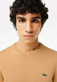 Maglione a maglia beige con collo a giro, che presenta un piccolo logo verde a forma di coccodrillo sul lato sinistro del petto. Texture liscia e design aderente.
