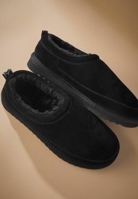 Czarne buty wsuwane wykonane z zamszu, z miękkim wyściółką i grubą gumową podeszwą. Posiadają teksturowany uchwyt z tyłu, ułatwiający zakładanie i zdejmowanie.
