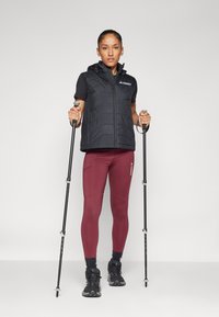 Svart isolerad väst med huva, svart kortärmad skjorta, vinröda leggings och vandringsstavar. Skorna är svarta med detaljerad snörning.