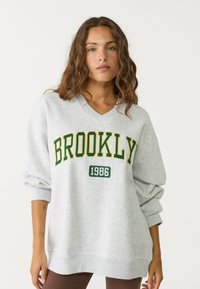 Ljusgrå sweatshirt med V-ringning, med "BROOKLYN" i gröna och gula bokstäver och året "1986" under. Lös passform med långa ärmar.