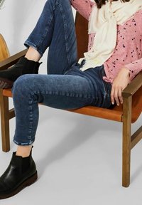 Jeans de mezclilla en un lavado medio, con los puños enrollados, combinados con botas de tobillo negras. La modelo lleva una blusa de patrón rosa y una bufanda crema, sentada en una silla de madera.