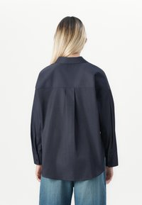 Woman with long blonde hair wearing a loose-fitting dark navy shirt and blue jeans, facing away against a plain white background.