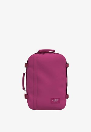 Mochila de viaje magenta con una textura suave, que cuenta con un bolsillo frontal con cremallera, un asa en la parte superior y correas de compresión laterales. Logo de "CabinZero" exhibido.