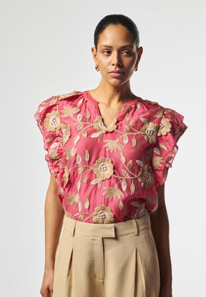 Frau mit dunklen Haaren in einem rosa Oberteil mit floraler Stickerei und Rüschenärmeln, trägt beige, hoch taillierte, plissierte Hosen und goldene Creolen.