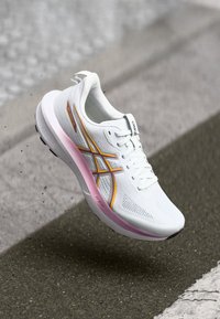 Weißer Sportschuh mit strukturiertem Obermaterial, orangefarbenen und pinken Akzenten, abgerundeter Zehenform und gepolsterter Sohle. Leichtes, elegantes und aerodynamisches Design.