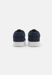 Marinblå mockasko med vit gummisula, med rundad form, texturerad ovandel och minimalistisk design utan synlig branding.