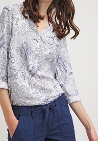 Blusa bianca con motivo paisley blu, scollo a V e maniche arrotolate, abbinata a pantaloni blu scuro con vita regolabile.