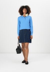 Lysblå langærmet sweater, denim-nederdel, sort håndtaske og strukturerede sorte sko. Blomstret accessory om halsen tilføjer detaljer.