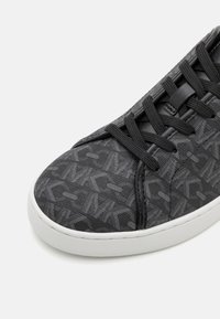 Zapatilla deportiva negra con parte superior de tela texturizada que presenta un patrón geométrico, puntera redonda y suela de goma blanca contrastante.